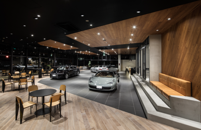 モダンで高級感のある自動車ショールームの店内。床は明るい木目調とダークグレーのタイルでデザインされ、展示スペースにはシルバーのスポーツカーや黒いセダンなどが並んでいる。手前にはカフェのようなテーブルと椅子のセットが置かれ、壁際にはオレンジ色のベンチシートが設置されている。黒い天井と木製パネル、そしてスポットライトが洗練された空間を演出している。